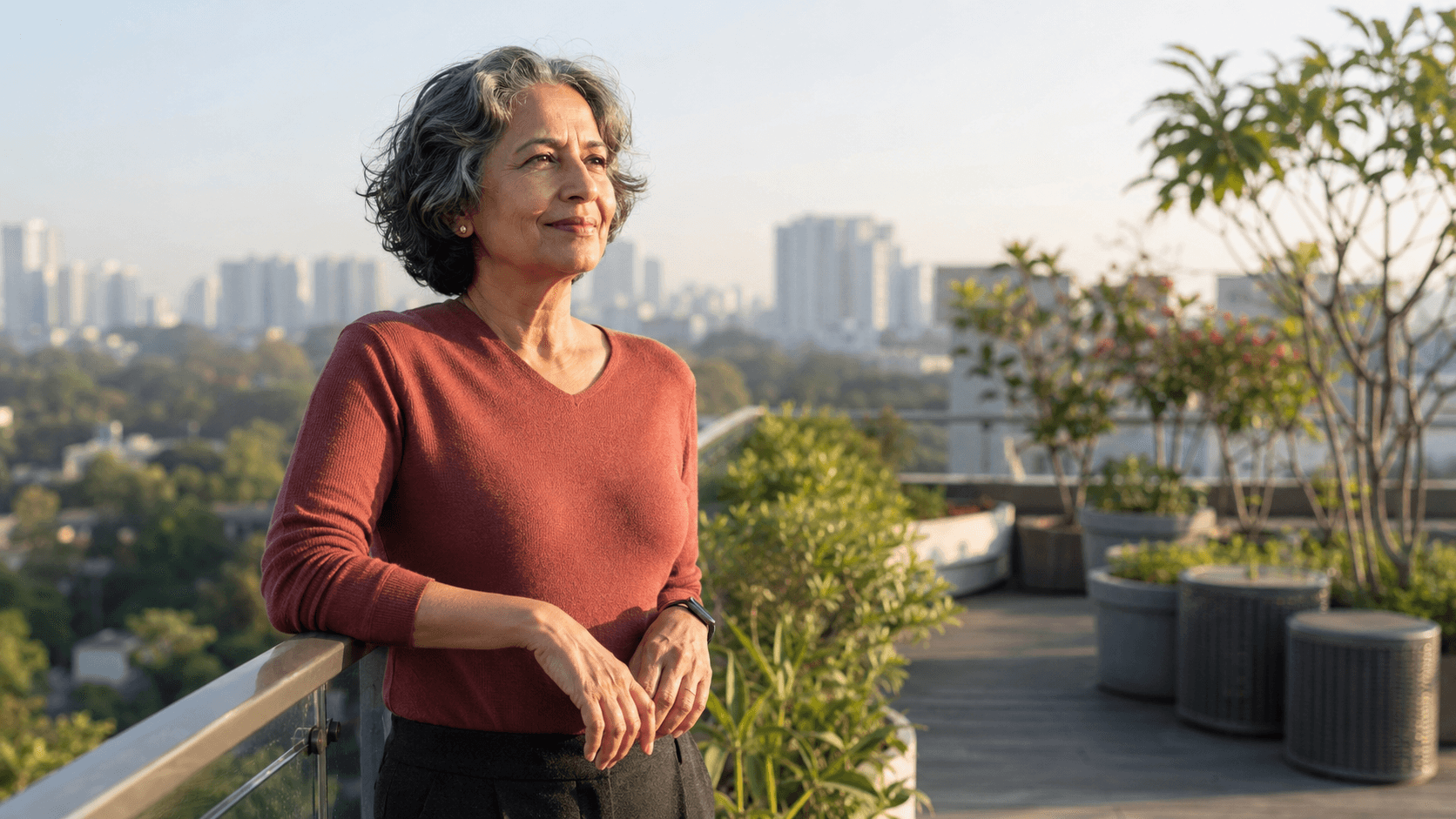 A confident Indian woman with natural silver hair standing on a sunlit rooftop garden overlooking the city